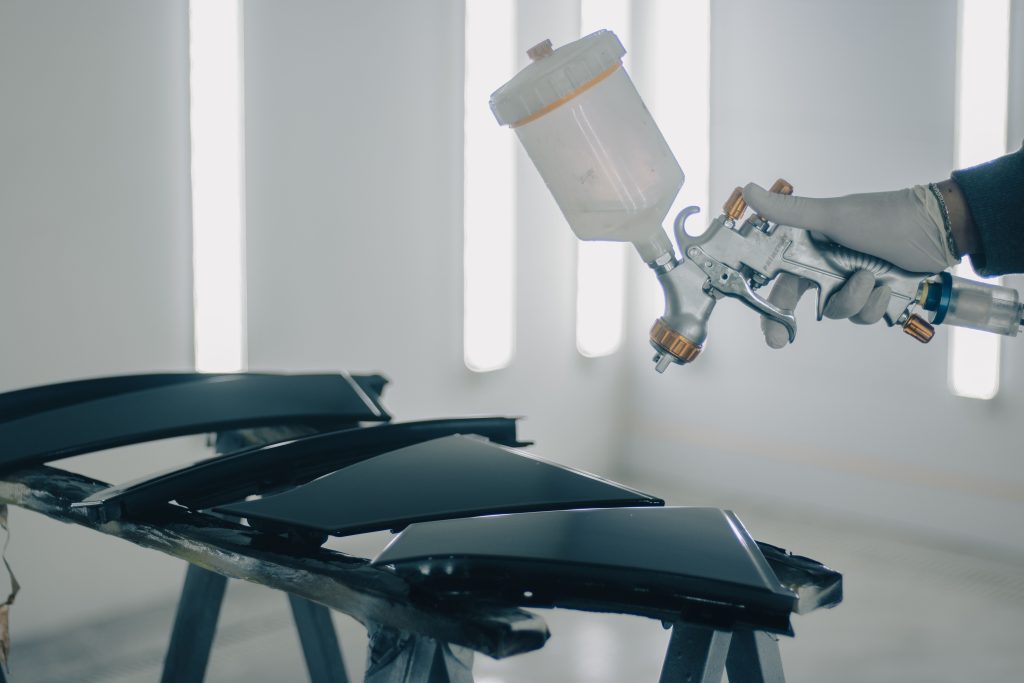 Automotive technician holding a professional paint spray gun near refinished vehicle body panels, representing Vehicle diagnostic system financing, EV repair technology financing, vehicle inspection equipment financing, and mechanic business financing options for automotive service businesses.